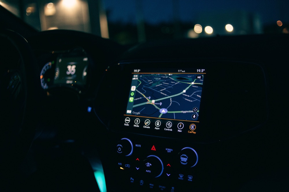 A car dashboard at night with a glowing in-car navigation map showing live traffic on nearby roads.