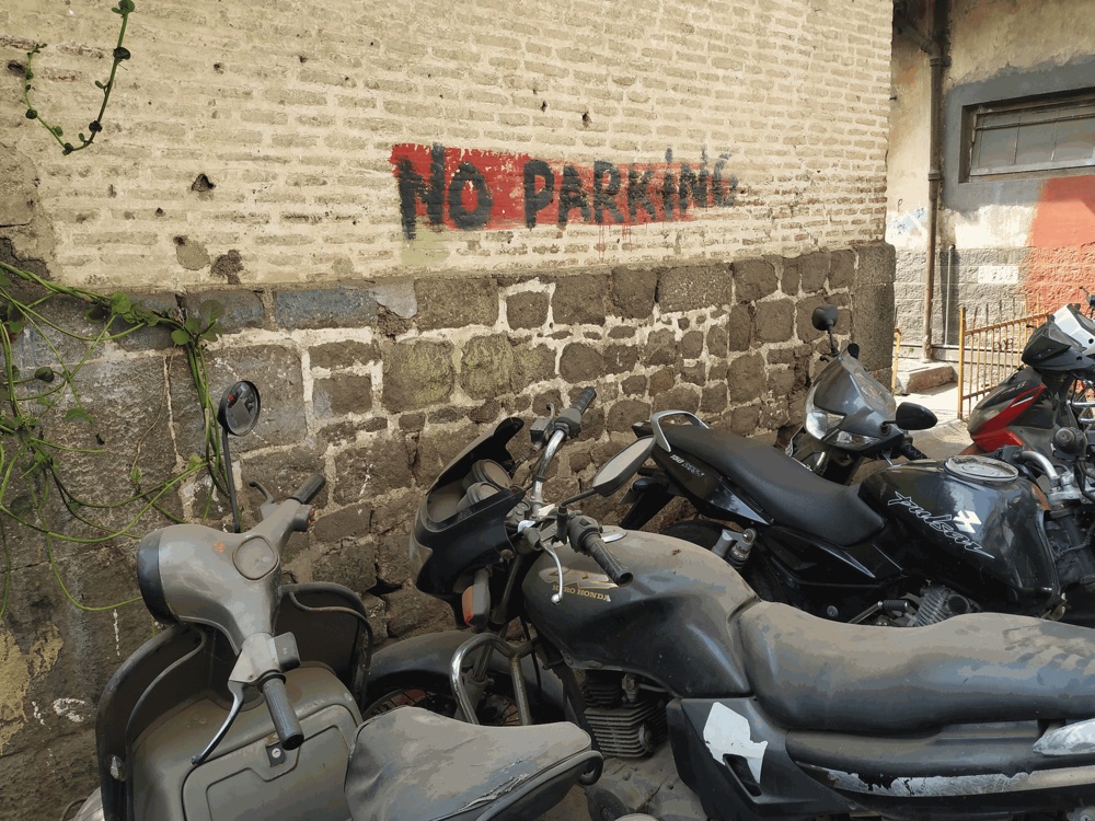 Dusty motorbikes and a scooter parked tightly against a stone wall, directly beneath a hand-painted NO PARKING sign.