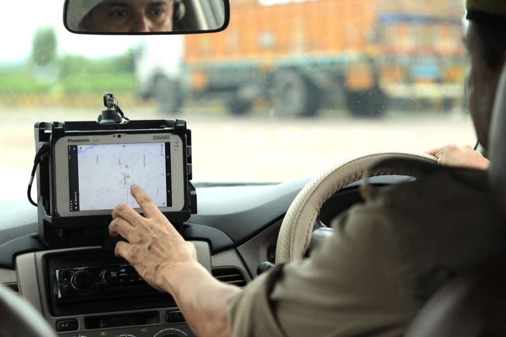 Inside a car, a uniformed driver taps a map on a rugged dashboard-mounted tablet, with a truck blurred beyond the windshield.